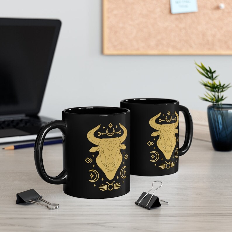 May include: Two black ceramic mugs with gold designs. The mugs feature a bull's head with celestial symbols and a crescent moon.