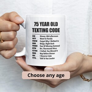 Può includere: Una tazza in ceramica bianca con il testo nero 75 YEAR OLD TEXTING CODE seguito da un elenco di definizioni umoristiche per acronimi comuni come DM, WTF, FWIW, BRB, FOMO, BTW, ICYMI, NBD, IDC e TTYL. La tazza ha un manico sul lato sinistro.
