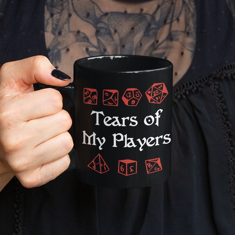 以下が含まれることがあります： 黒いコーヒーカップに、白い文字で「Tears of My Players」と書いてあり、その周りに赤いサイコロのグラフィックが描かれている。