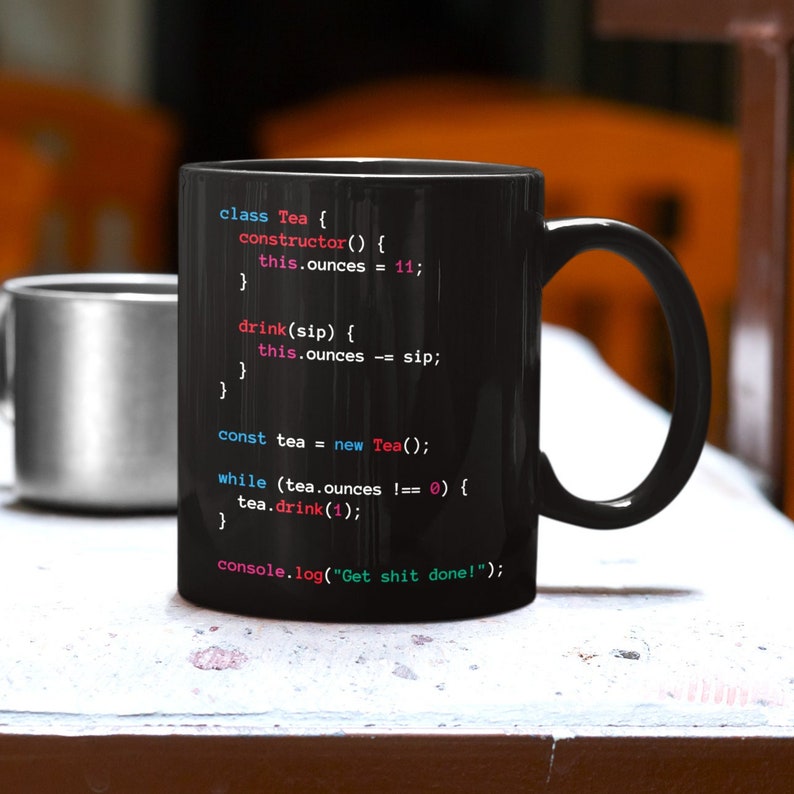 May include: Black ceramic mug featuring programming code, including "class Tea" and "console.log("Get shit done!")". The mug is on a white surface, with a silver mug visible in the background.