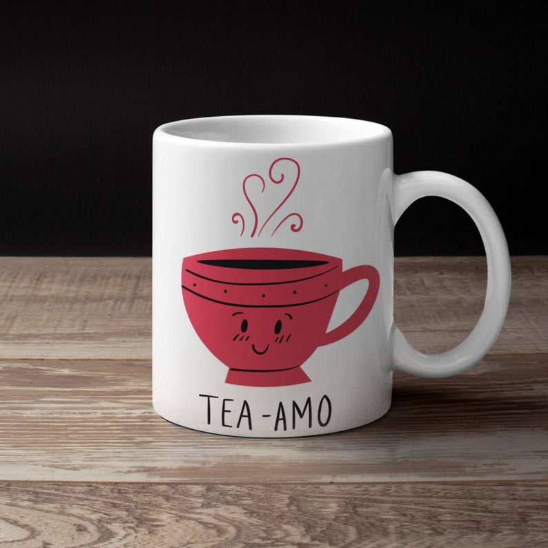 May include: White ceramic mug with a red teacup with a smiling face and steam coming out of it. The text "TEA-AMO" is printed below the teacup.