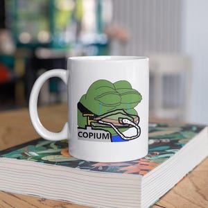 以下が含まれることがあります： 悲しい表情の緑色のカエルのキャラクターと「COPIUM」の文字が描かれた白いセラミックマグカップ。マグカップは、花柄のデザインの書籍の上に置かれています。マグカップには湾曲したハンドルが付いています。