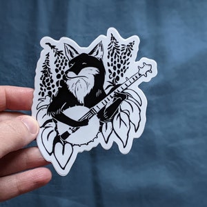 May include: Black and white sticker featuring a fox playing a banjo. The fox is surrounded by leaves and trees. The sticker has a white border and is on a blue background.