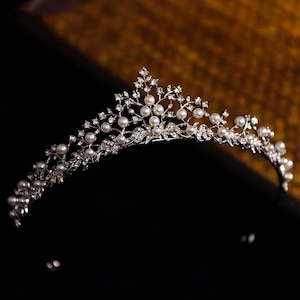 May include: A silver tiara with a floral design. The crown features faux pearls and clear crystals, arranged in a delicate pattern. The tiara is set against a black background with a blurred, golden-brown backdrop.
