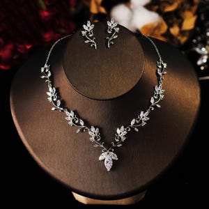 Könnte beinhalten: Ein silberfarbenes Halsketten- und Ohrring-Set, das auf einer braunen Schmuckbüste präsentiert wird. Das Set hat ein zartes Blumendesign mit klaren, funkelnden Edelsteinen. Die Halskette hat einen Anhänger mit einer Ansammlung von Steinen.