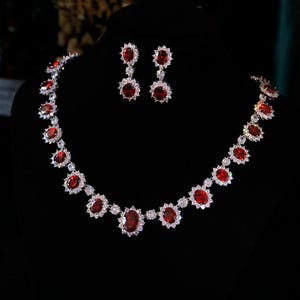 May include: A necklace and earring set with oval, deep red gemstones and clear surrounding stones. The jewellery is set in silver-toned metal. The necklace and earrings are displayed against a black background, creating a striking contrast.