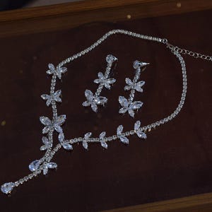 May include: A silver-toned necklace and earring set. The necklace features a floral design with clear, sparkling stones. The earrings mirror the floral motif. The jewelry set is displayed on a dark brown surface.