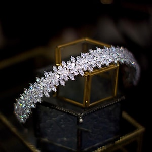 Puede incluir: Una tiara plateada con un diseño floral, adornada con numerosos cristales transparentes. La tiara se exhibe sobre una pequeña caja de cristal con marco dorado, sobre un fondo oscuro. Los cristales capturan la luz, creando un efecto deslumbrante.