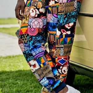 May include: Jogger pants featuring a vibrant patchwork design with various geometric and abstract patterns in shades of blue, orange, yellow, purple, and black. The pants have elasticized cuffs at the ankles and are paired with white sneakers.