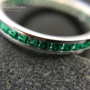 May include: A close-up of a silver band with a row of emerald green gemstones set in the metal. The band is on a grey textured surface.