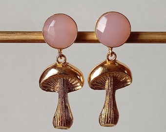 May include: A pair of gold mushroom earrings with pink gemstone tops. The earrings are a delicate and whimsical design.