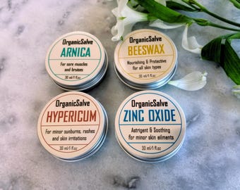 Puede incluir: Cuatro pequeños botes redondos de metal de OrganicSalve, cada uno etiquetado con un ingrediente natural diferente: Árnica, Cera de Abejas, Hipérico y Óxido de Zinc. Las etiquetas incluyen el nombre del producto y una breve descripción de su uso. Cada bote contiene 30 ml / 1 fl oz.