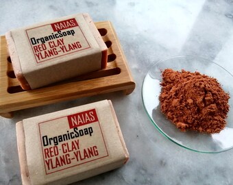 May include: Two bars of handmade soap on a wooden soap dish. The soap is labeled "NAIAS Organic Soap Red Clay Ylang-Ylang". A small dish of red clay powder is in the background.
