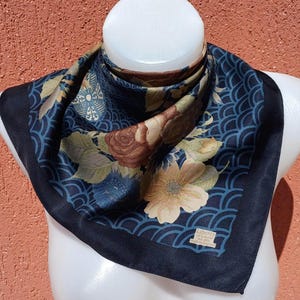May include: A square silk scarf with a floral pattern in shades of brown, beige, and blue. The scarf has a dark blue border and a repeating wave-like design. The label reads "Liberty of London, ALL SILK, MADE IN ENGLAND."