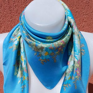 May include: A vibrant blue silk scarf with a floral pattern. The scarf features a square shape with a bright blue background and a border of flowers in shades of yellow, pink, and green. The scarf is draped over a white mannequin.