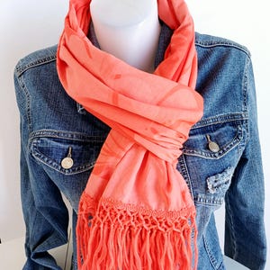 May include: Coral-colored scarf with a fringed edge, draped over a denim jacket. The scarf has a subtle pattern and is wrapped around a white mannequin head. The denim jacket has silver buttons and pockets.