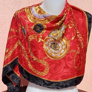 Op de afbeelding: Een rode zijden sjaal met een gouden ketting- en horlogedesign. De sjaal heeft een zwarte rand en is gedrapeerd over een witte mannequinhoofd. Het ontwerp van de sjaal omvat gouden kettingen, horloges en andere sierlijke details.