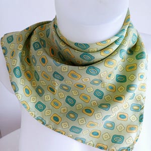 May include: A square scarf with a repeating pattern of geometric shapes in shades of green, yellow, and teal. The scarf is draped around a white mannequin head, showcasing its design and texture. The scarf appears to be made of a lightweight fabric.