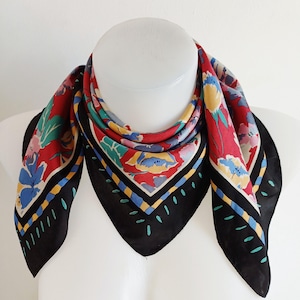 May include: A square scarf with a floral pattern. The scarf features a red background with colorful flowers in blue, yellow, and pink. The border is black with a geometric design. The scarf is displayed on a white mannequin.
