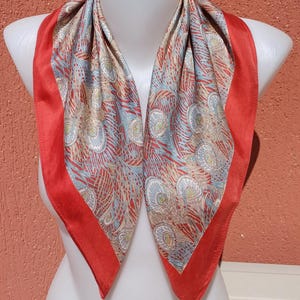 May include: A silk scarf with a vibrant peacock feather pattern in shades of blue, beige, and red. The scarf has a solid red border and is displayed on a white mannequin.
