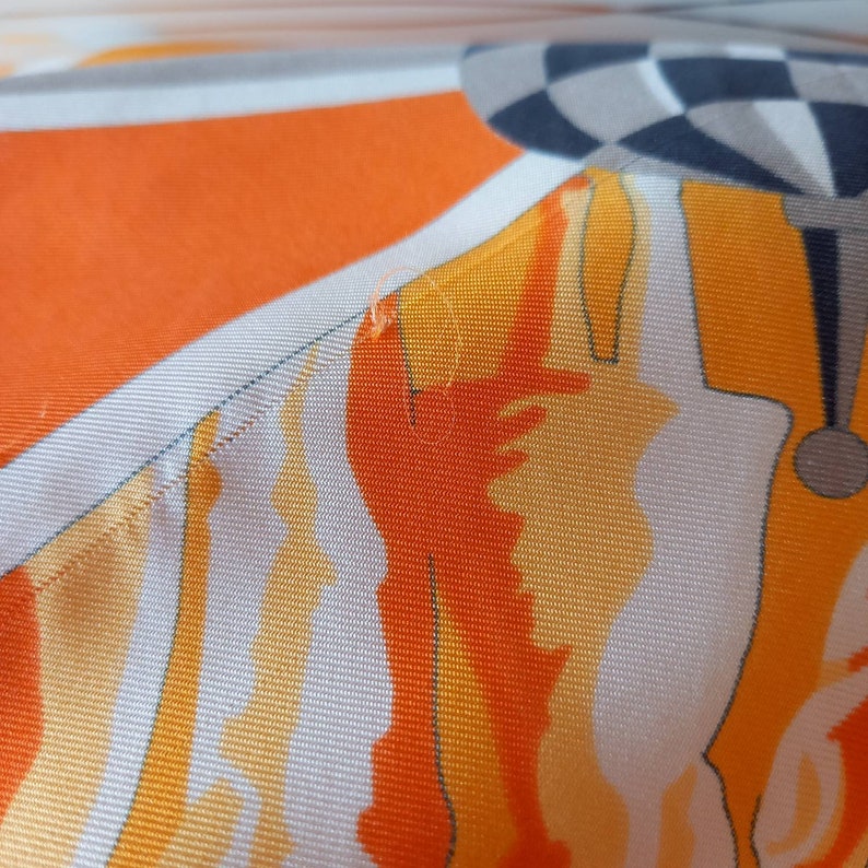 May include: Close-up of a white fabric with orange and yellow abstract patterns. The fabric has a subtle texture and a slightly faded appearance.