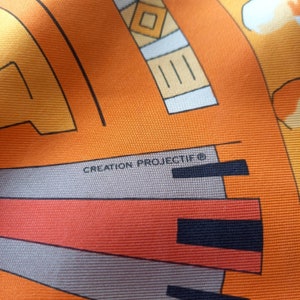 May include: Close-up of a fabric with a geometric pattern in orange, yellow, and gray. The text "CREATION PROJECTIF&reg;" is visible in the lower left corner.