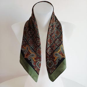 May include: A patterned scarf with a repeating design of red, brown, yellow, and green. The scarf is displayed on a white mannequin. The edges of the scarf are a solid green color. The scarf is made of a silky material.