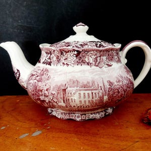Könnte beinhalten: Eine Vintage-Teekanne aus Keramik mit einem bordeauxroten und weißen Transferdruck-Design. Die Teekanne zeigt eine Landschaftsszene mit einem Gebäude und floralen Mustern. Sie hat einen gebogenen Ausguss, einen Henkel und einen Deckel mit dekorativem Knauf. Die Teekanne steht auf einer Holzoberfläche.