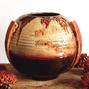 Könnte beinhalten: Eine handgefertigte Keramikvase in Kugelform mit Farbverlauf von hellbeige zu tiefbraun. Die Vase hat zwei orange-braune Griffe und steht auf einer Holzkiste mit verblasstem Text. Getrocknete rote Blumen sind um die Vase angeordnet. Der Durchmesser beträgt ca. 20 cm.