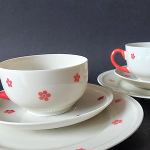 Könnte beinhalten: Zwei weiße Teetassen aus Keramik mit passenden Untertassen, verziert mit einem wiederkehrenden Muster aus kleinen orangefarbenen Blumen mit blauen Zentren. Jede Tasse hat einen leuchtend orangefarbenen Henkel und einen dezenten Perlenrand.