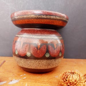 May include: A two-piece ceramic vase with a bulbous base and a smaller, flared top. The vase features a brown, orange, and red color scheme with a floral pattern around the base. The vase is sitting on a wooden surface.
