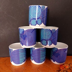 May include: A set of six white ceramic cups stacked in a pyramid formation. Each cup features a repeating graphic pattern of concentric circles and horizontal lines in shades of deep royal blue, navy, and bright turquoise against a white background.
