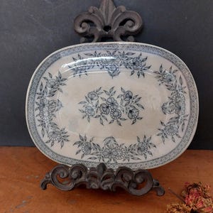 Może przedstawiać: Owalny ceramiczny półmisek z kwiatowym wzorem w odcieniach niebieskiego i białego. Półmisek ma dekoracyjne obramowanie i jest podparty ciemnym metalowym stojakiem. Wzór kwiatowy skoncentrowany jest na środku i na krawędziach.