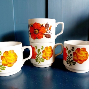 Può includere: Quattro tazze in ceramica bianca con disegni floreali arancioni, gialli e verdi. Le tazze hanno una forma classica con manico e sono impilate e disposte su una superficie blu. Il motivo floreale si ripete su ogni tazza.