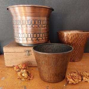 May include: Three copper planters of varying sizes. The largest planter has a brick-like pattern, while the medium-sized one has a textured surface. The smallest planter is smooth. Dried flowers and a wooden box are also present.