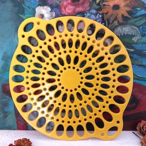 Könnte beinhalten: Ein leuchtend gelbes, kreisförmiges Küchensieb mit einem komplizierten Muster aus ovalen und runden Löchern. Das Sieb hat zwei gebogene Griffe und eine zentrale Nabe. Der Hintergrund zeigt ein farbenfrohes Blumenbild.