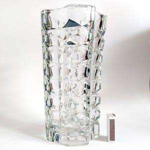 Può includere: Un vaso di vetro trasparente e sfaccettato con forma rettangolare e superficie strutturata. Il vaso è alto circa 25 cm. Un fiammifero è accanto al vaso per la scala.