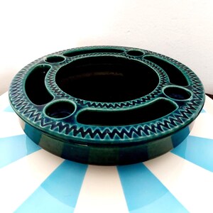 Può includere: Un posacenere rotondo in ceramica color verde acqua, con una vasca centrale e molteplici aperture circolari e oblunghe sul perimetro. Il bordo presenta un motivo a zig-zag. Il posacenere è su una superficie a strisce blu e bianche.