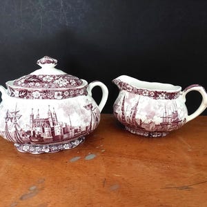 Può includere: Una zuccheriera con coperchio e una lattiera abbinata. Entrambi i pezzi in porcellana bianca presentano un motivo transferware bordeaux con un paesaggio urbano e bordi floreali. La zuccheriera ha due manici.