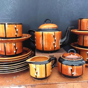 Könnte beinhalten: Vintage Teeservice in Orange und Schwarz. Das Set umfasst eine Teekanne, Tassen, Untertassen, einen Sahnekännchen und eine Zuckerdose. Jedes Stück ist mit vertikalen schwarzen Streifen und weißen floralen Akzenten verziert.