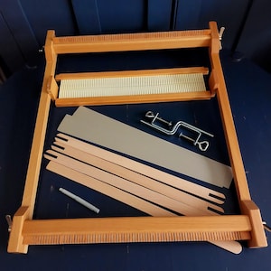 Peut inclure: Un cadre de métier à tisser en bois avec un peigne supérieur et inférieur, accompagné d'un peigne séparé, quatre navettes plates en bois, un outil en plastique gris et deux serre-joints en métal. Les composants sont disposés pour l'assemblage.