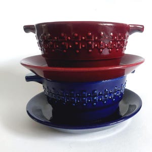 Könnte beinhalten: Gestapelte Keramikschalen und Untertassen in Rot- und Blautönen. Die Schalen haben Griffe und ein strukturiertes, geometrisches Muster. Die Untertassen sind einfarbig und ergänzen die Schalen. Ein stilvolles Set zum Servieren.