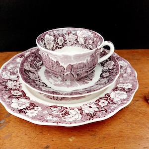 Puede incluir: Juego de té vintage de tres piezas en morado y blanco. El juego incluye una taza, un platillo y un plato, todos con un patrón floral y paisajístico. Los bordes del plato y el platillo son festoneados. La taza está colocada sobre el platillo, que está sobre el plato.