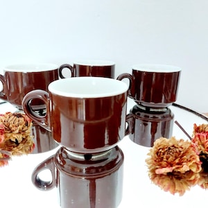 Pode incluir: Quatro xícaras de café de cerâmica marrom com interiores brancos e alças arredondadas são dispostas em uma superfície reflexiva. Flores secas estão espalhadas ao redor das xícaras. As xícaras são de uma cor marrom escura rica.