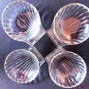 Op de afbeelding: Vier heldere, schelpvormige glazen schalen in een cirkelvormig patroon. Elke schaal heeft een geribbeld ontwerp en een kleine rechthoekige basis met tekst. De schalen staan op een donkere ondergrond.