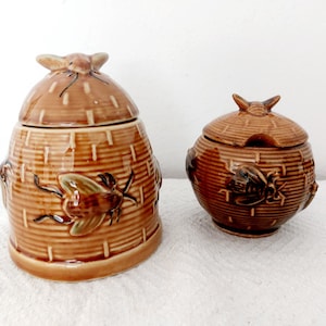 May include: Two brown ceramic containers with lids. The larger one is shaped like a beehive, and the smaller one is round. Both have a bee design and a textured surface.