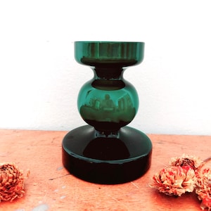 May include: A dark emerald green glass candle holder featuring a wide circular base, a rounded bulbous centre section, and a cylindrical top opening. The smooth, reflective surface of the glass catches light, creating a deep translucent colour.