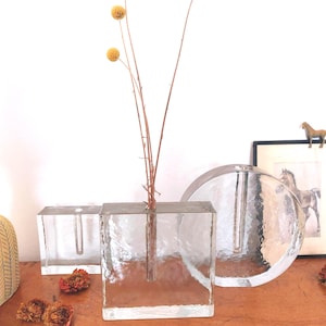 Könnte beinhalten: Klare Glasvasen in rechteckiger und kreisförmiger Form. Eine Vase enthält getrocknete gelbe, kugelförmige Blumen. Die Vasen stehen auf einer Holzoberfläche mit einem gerahmten Pferdedruck und einer kleinen Pferdfigur im Hintergrund.