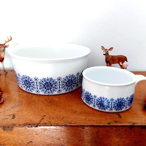Op de afbeelding: Twee witte keramische kommen met een blauw sneeuwvlokpatroon aan de basis. Een grotere kom en een kleinere kom met een handvat worden gepresenteerd op een houten oppervlak. Decoratieve hertenbeeldjes zijn ook aanwezig.