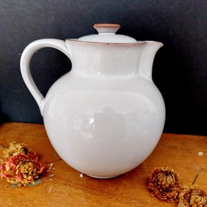 Könnte beinhalten: Ein Set aus drei Keramikbechern mit cremefarbener, gesprenkelter Glasur. Jeder Becher zeigt ein handgemaltes Design: ein blau-grünes Blatt, eine gelbe Blume und eine rote Beere. Die Becher haben ein schlichtes, modernes Design.